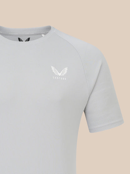 A close-up of a light gray short-sleeve athletic t-shirt with a white winged logo and the brand name 'CASTORE' printed on the left chest, set against a solid beige background.