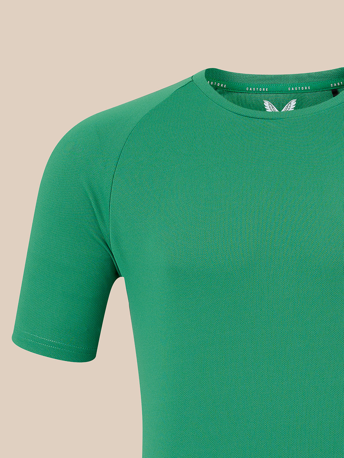 A close-up of a green short-sleeve athletic t-shirt on a solid beige background, showing the collar with a white wing logo and the brand name 'CASTORE' printed inside.