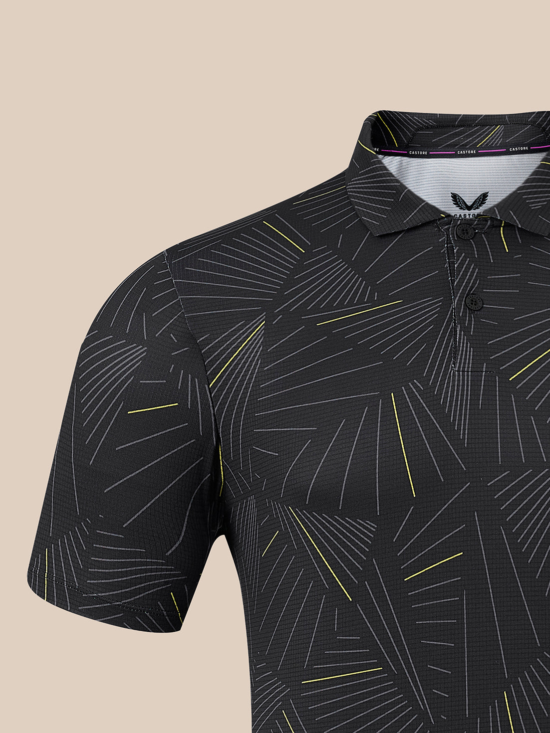 A close-up of a short-sleeve polo shirt with a black base and an abstract geometric line pattern in white and yellow, shown against a solid beige background.