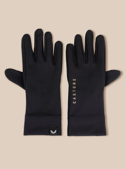 A pair of black long gloves laid flat on a light beige background. One glove shows the back with a small black logo tab near the cuff, and the other shows the front with the vertical brand name 'CASTORE' printed in white.
