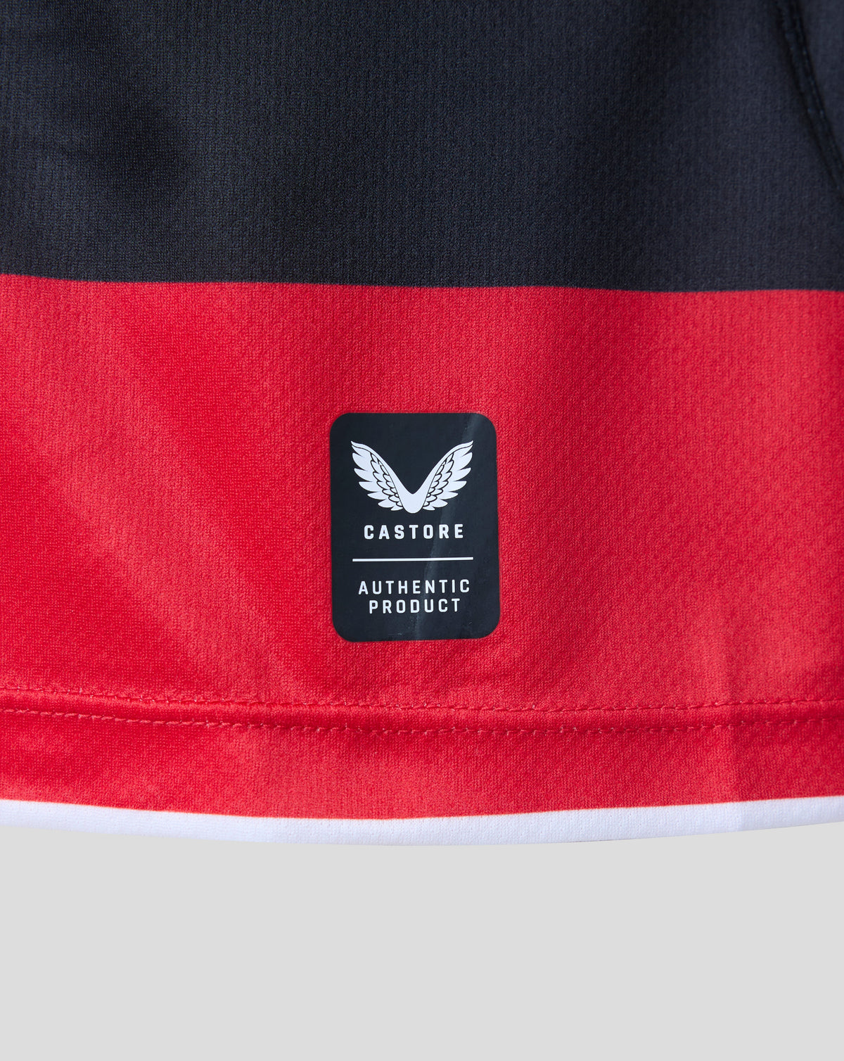 Close-up of a red and black athletic garment showing a black Castore brand patch with a white wing logo and 'AUTHENTIC PRODUCT' text.