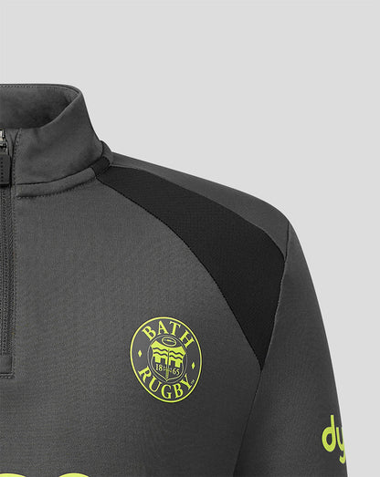 Close-up of a dark gray athletic quarter-zip pullover with a black shoulder panel, featuring a yellow circular Bath Rugby crest on the chest and a yellow logo on the sleeve, set against a light gray background.