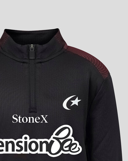Close-up of a black athletic quarter-zip pullover with a mock neck, featuring a white crescent and star logo on the chest, 'StoneX' text, and a large white sponsor logo across the lower front. The shoulders have subtle burgundy textured panels.
