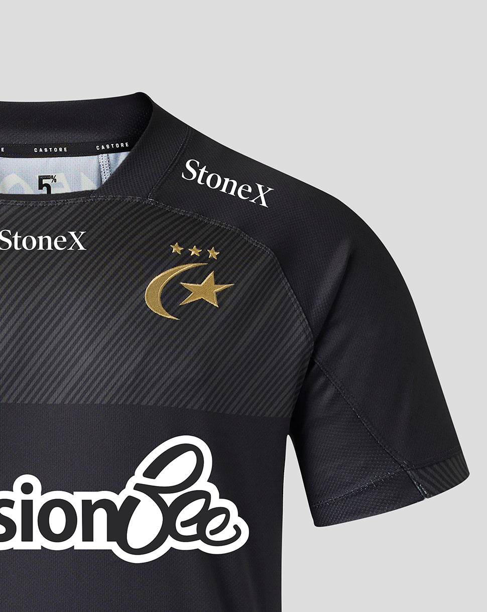 Close-up of a black short-sleeve sports jersey with diagonal textured paneling on the chest, a gold crescent and star logo with three stars, and sponsor logos including StoneX and PensionBee on a light gray background.