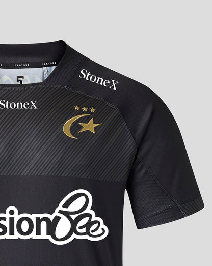 Close-up of a black short-sleeve sports jersey with diagonal textured paneling on the chest, a gold crescent and star logo with three stars, and sponsor logos including StoneX and PensionBee on a light gray background.