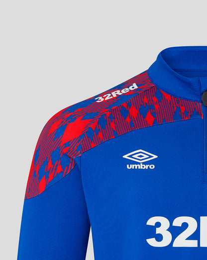 Close-up of a blue and red patterned athletic quarter-zip jacket by Umbro, featuring the white Umbro logo and '32Red' text on the shoulder, set against a light gray background.