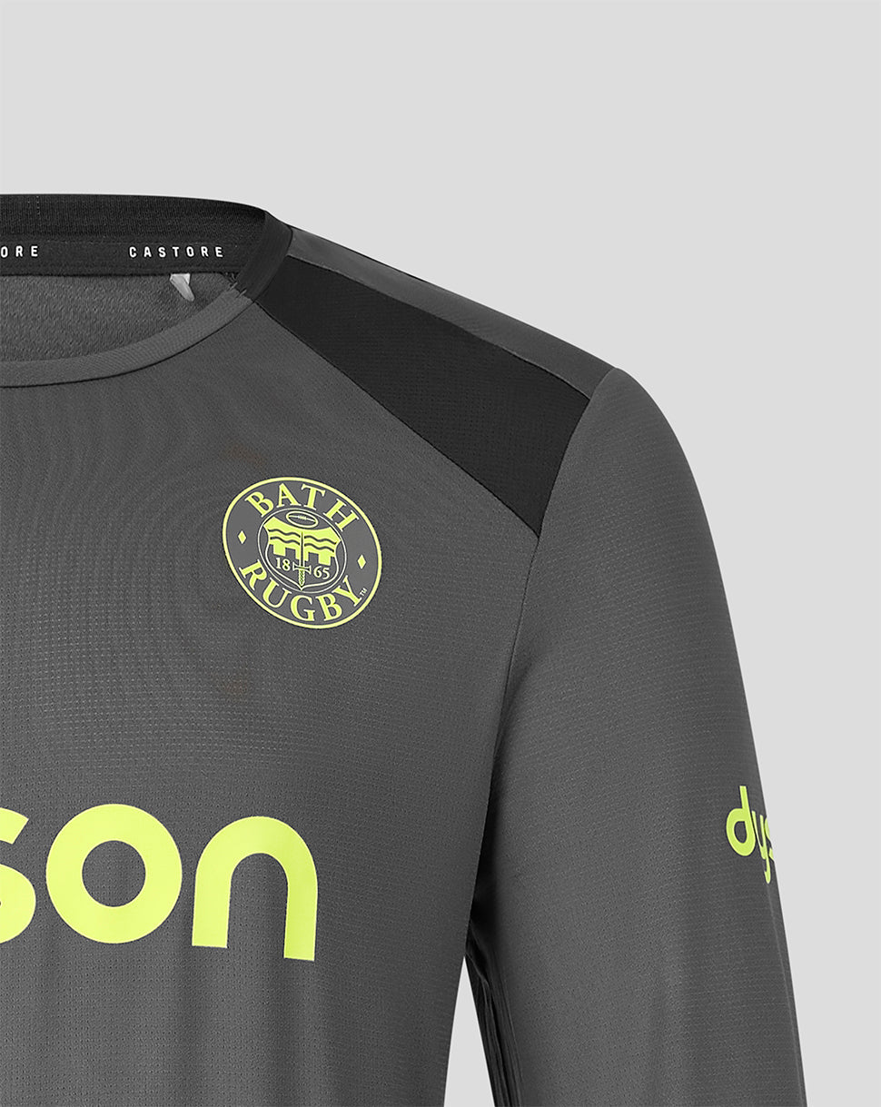 Close-up of a dark gray long-sleeve sports jersey with a black shoulder panel, a circular Bath Rugby crest on the chest, and neon yellow-green branding details on a light gray background.