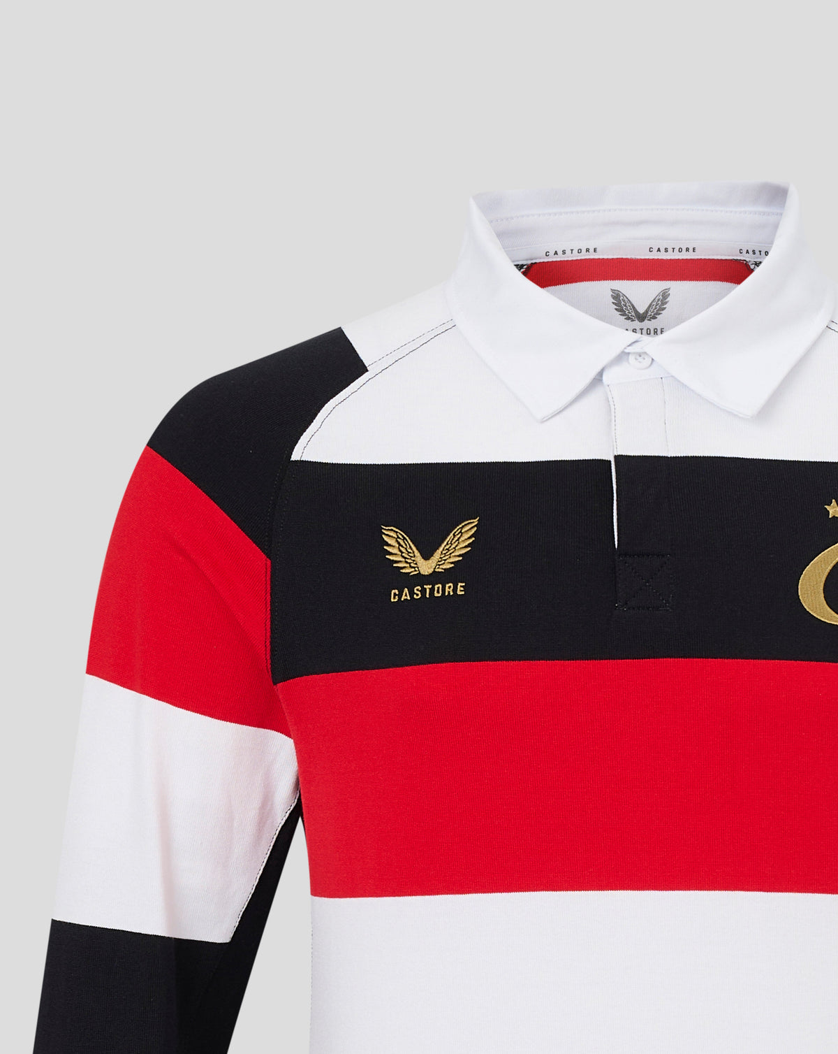 A close-up of a Castore long-sleeve rugby shirt featuring horizontal color-blocked stripes of white, black, and red, with gold embroidered logos on the chest.