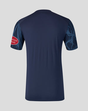 A navy blue short-sleeve athletic jersey shown from the back, featuring a red oval logo on the right sleeve and a tonal dark blue graphic pattern on the left sleeve, set against a solid light gray background.