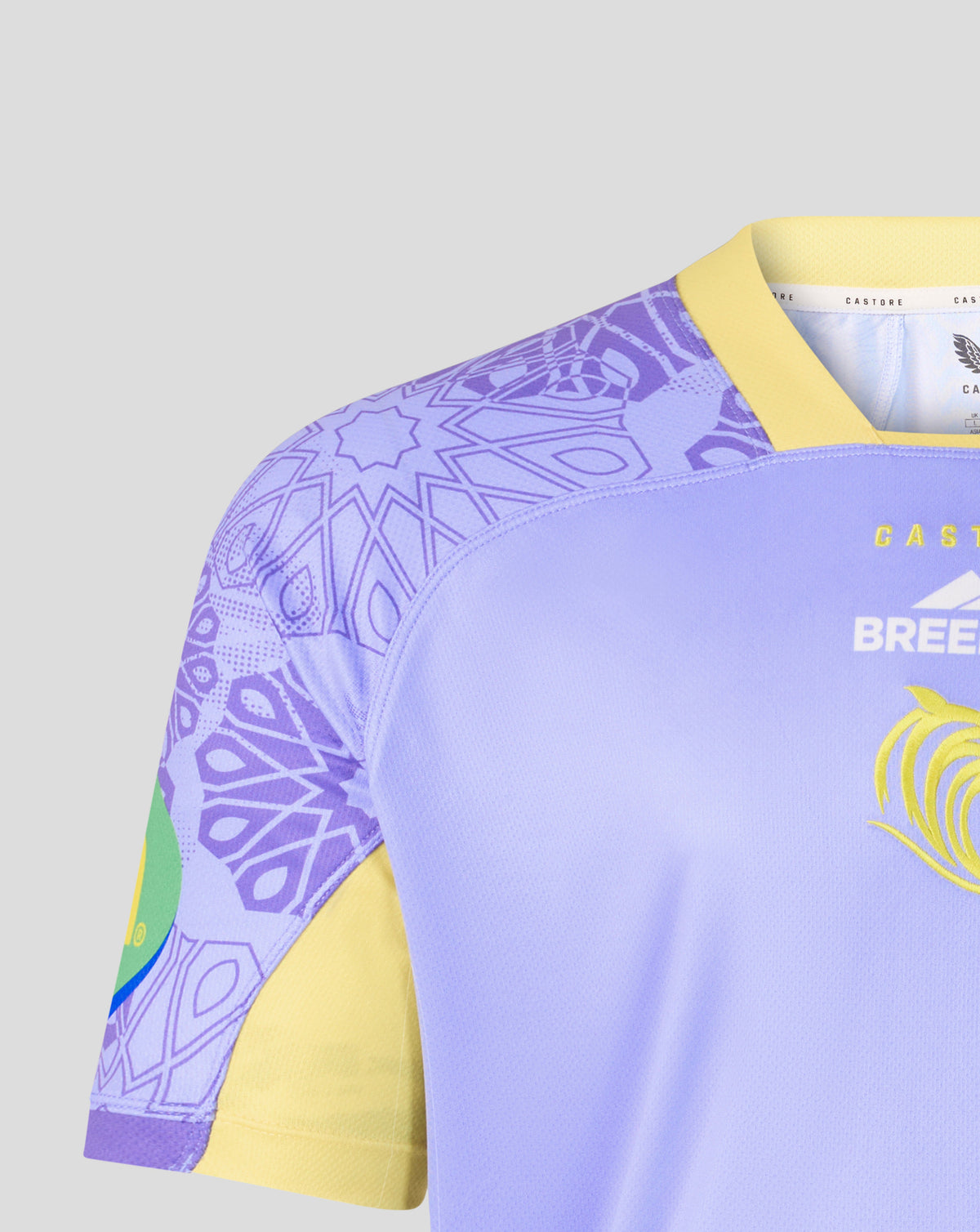 Close-up of a soccer jersey showing the shoulder and upper chest area. The jersey has a light purple body, a yellow V-neck collar and sleeve cuffs, a purple geometric pattern on the shoulder, and a yellow graphic on the chest.