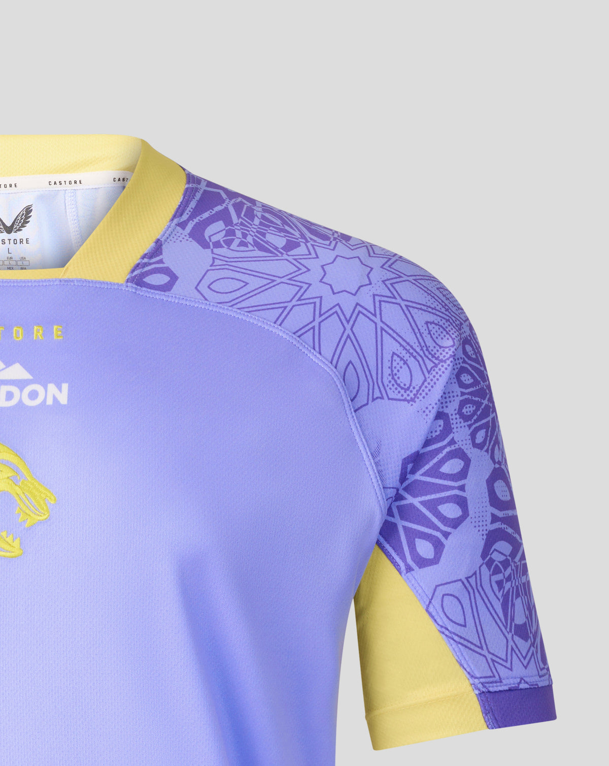 Close-up of a soccer jersey showing the shoulder and sleeve area. The jersey is light purple with a yellow collar and sleeve cuff, featuring a purple geometric pattern on the upper sleeve and shoulder, and yellow accents on the side.