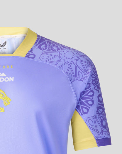 Close-up of a rugby jersey showing the shoulder and sleeve area. The jersey is light purple with a yellow collar and sleeve cuff, featuring a purple geometric pattern on the upper sleeve and shoulder, and yellow accents on the side.