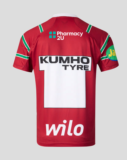 The back of a red sports jersey with green and white sleeve stripes, featuring prominent sponsor logos including Pharmacy 2U, Kumho Tyre, Wilo, and a JBL patch on a light gray background.