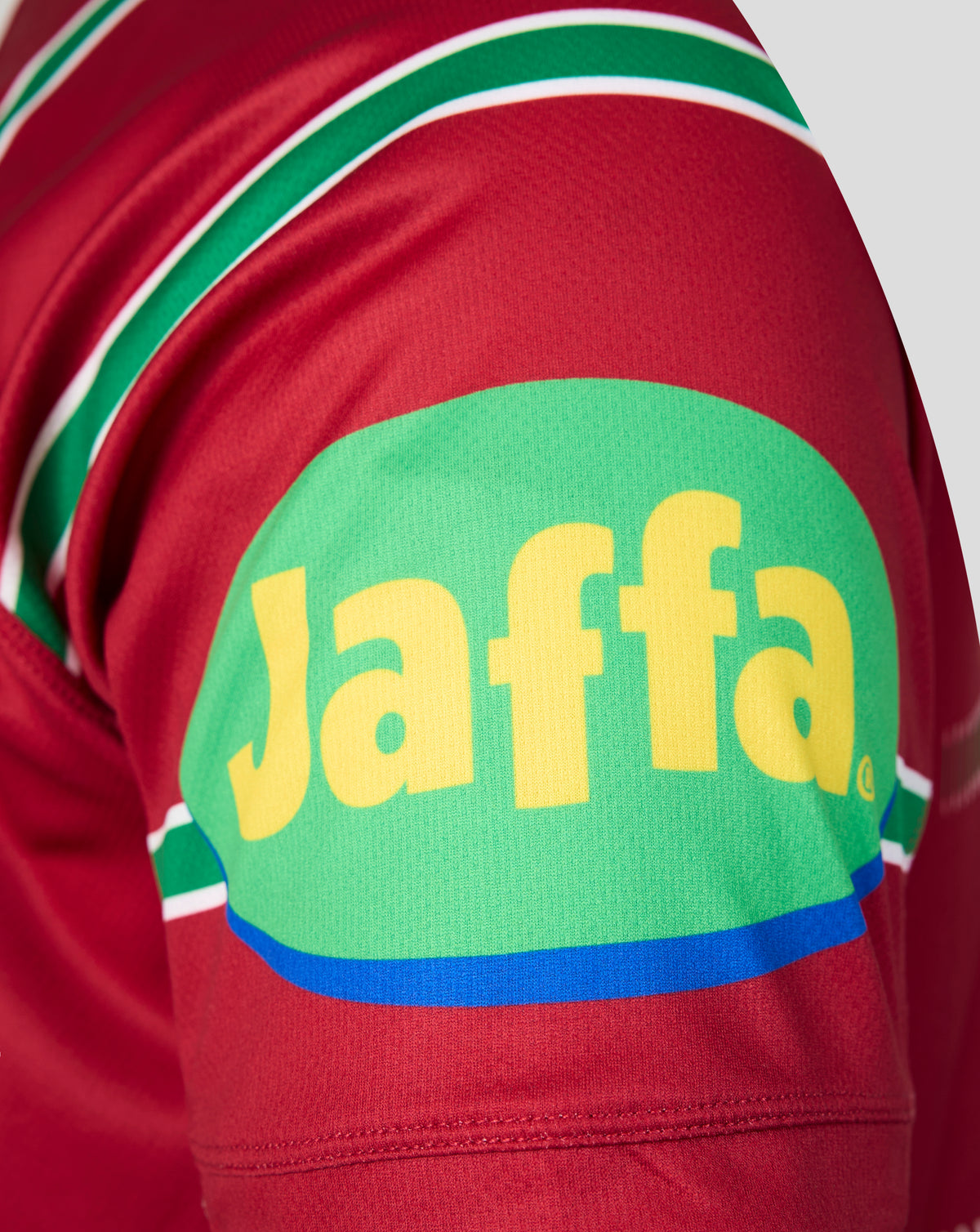 Close-up of a red athletic jersey sleeve featuring a large green oval patch with the yellow Jaffa logo and green, white, and blue stripe details.