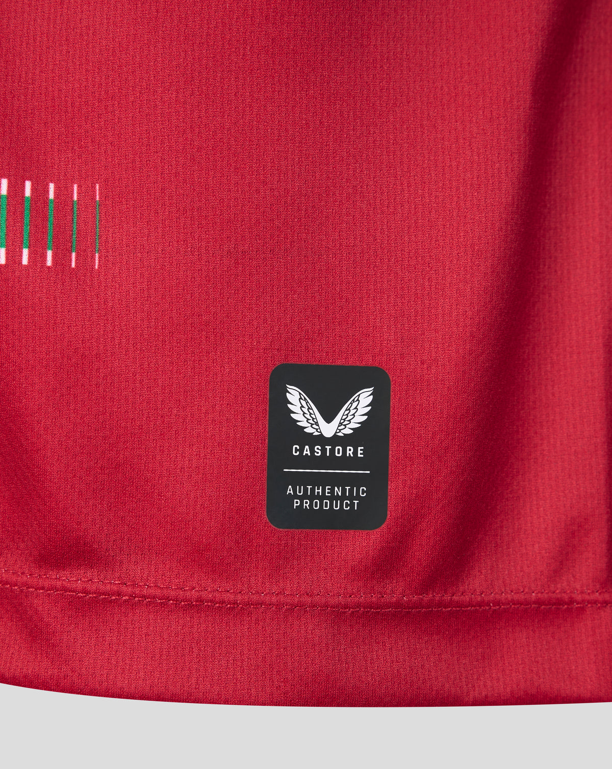 Close-up of a red athletic top showing a black Castore brand patch with white wing logo and 'AUTHENTIC PRODUCT' text, plus a small striped detail on the left side.