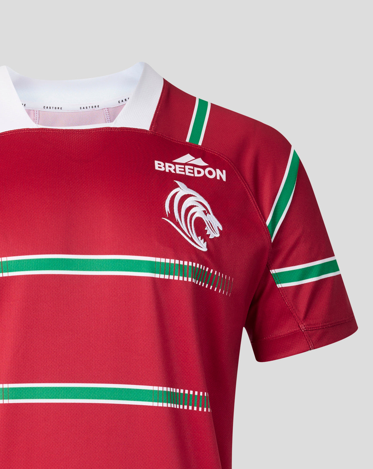 A close-up of a red short-sleeve sports jersey with green and white horizontal stripes, a white collar, and a white 'BREEDON' logo with a lion emblem on the chest, shown against a light gray background.