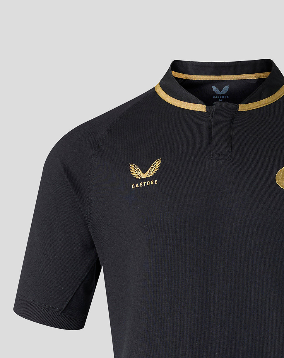 Close-up of a black short-sleeve polo shirt with gold trim on the collar, featuring a gold Castore wing logo on the left chest and a gold crest on the right chest, set against a light gray background.