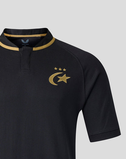 Close-up of a black short-sleeve polo shirt with gold trim on the collar and a gold crescent and star emblem with three stars on the chest, shown on a light gray background.