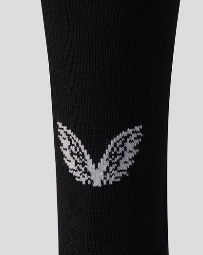 A close-up of a black knit sock featuring a white knitted butterfly motif on the lower calf, set against a solid light gray background.