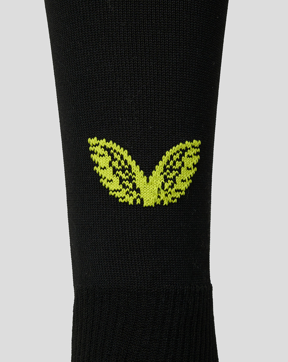 A close-up of a black knit sock featuring a bright yellow-green embroidered winged logo on the calf, set against a solid light gray background.