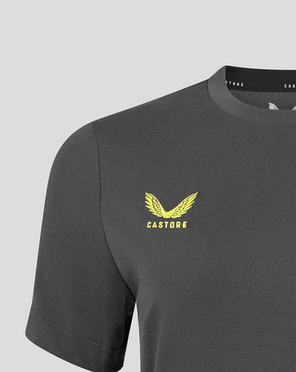 A close-up of a dark gray Castore short-sleeve t-shirt featuring a yellow winged eagle logo and the brand name 'CASTORE' printed on the left chest, set against a light gray background.