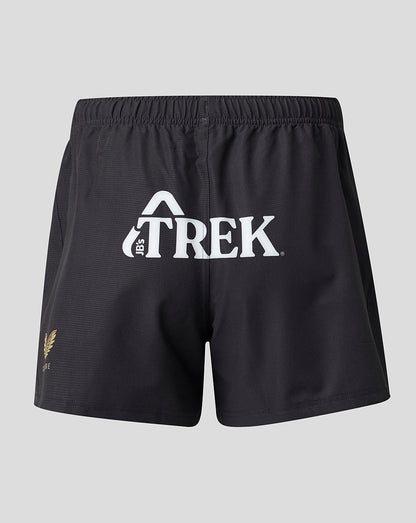 Black athletic shorts with a white 'TREK' logo on the front and a small gold fern logo on the lower left leg, shown against a light gray background.