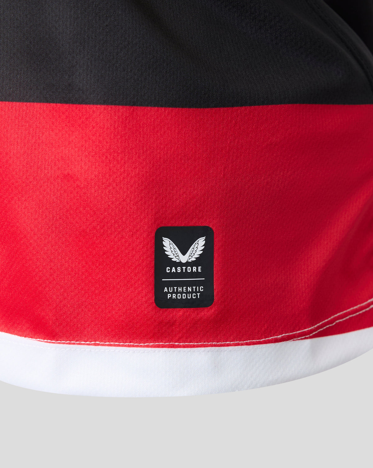 Close-up of a color-blocked athletic garment, likely shorts, featuring a wide red band across the middle, a black top section, and a white bottom hem. A black Castore 'Authentic Product' logo patch is sewn onto the red section.