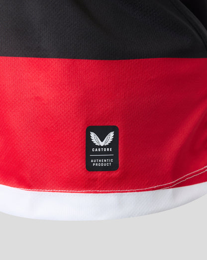 Close-up of a color-blocked athletic garment, likely shorts, featuring a wide red band across the middle, a black top section, and a white bottom hem. A black Castore 'Authentic Product' logo patch is sewn onto the red section.