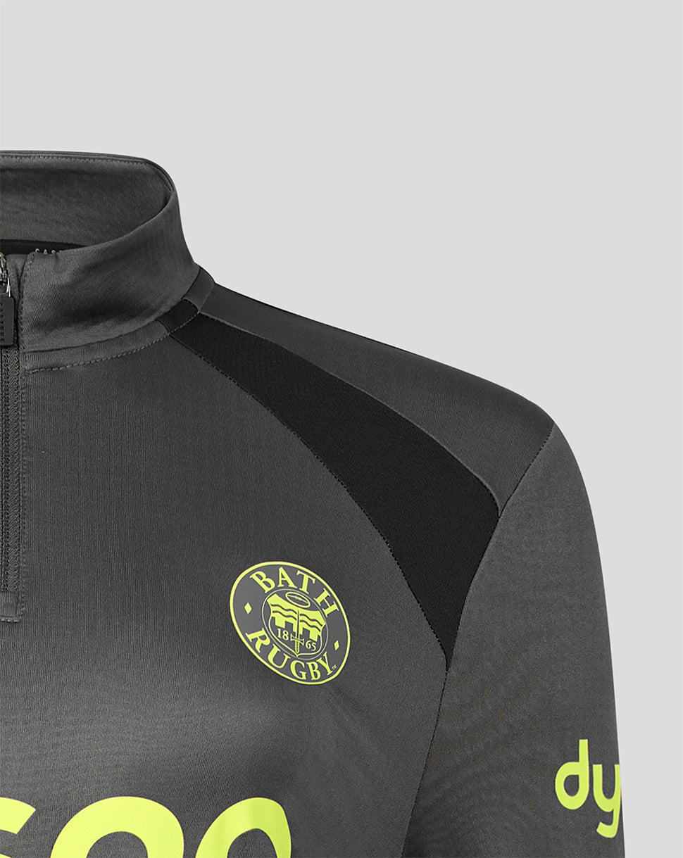 Close-up of a dark gray athletic quarter-zip top with a black collar and shoulder panel, featuring a yellow and black Bath Rugby crest on the chest and neon yellow branding on the sleeve, set against a solid gray background.