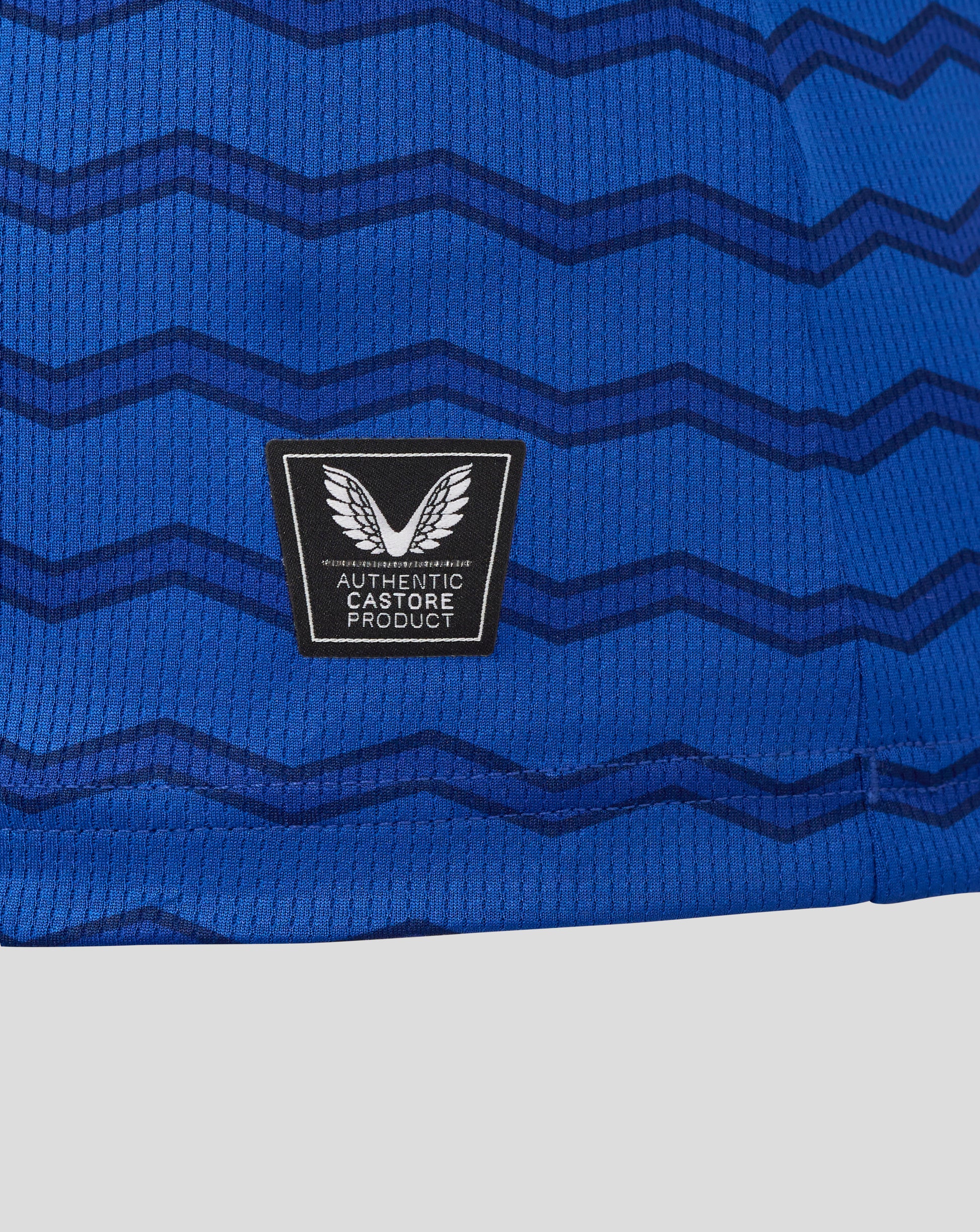 Close up of blue Everton women's 25/26 home shirt fabric with dark chevron waves and a black patch featuring white wings and Authentic Castore Product text.
