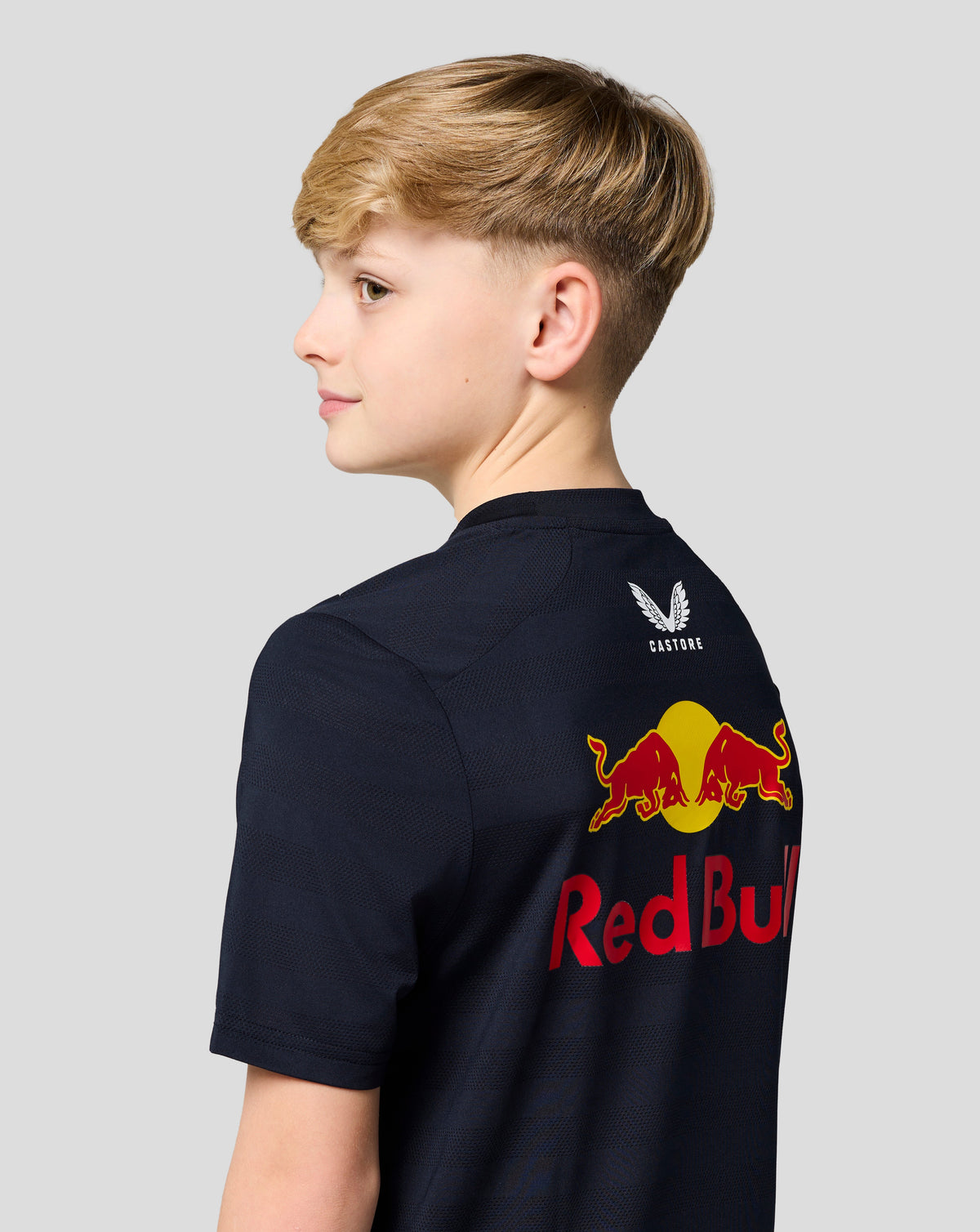 Side profile of a young boy wearing a navy Red Bull Racing T shirt with a white wing logo on the back