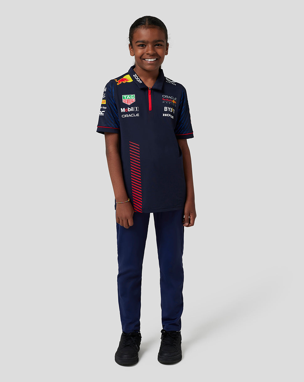 Young girl wearing a night sky navy Junior Oracle Red Bull Racing polo shirt with sponsor logos, full-body view.