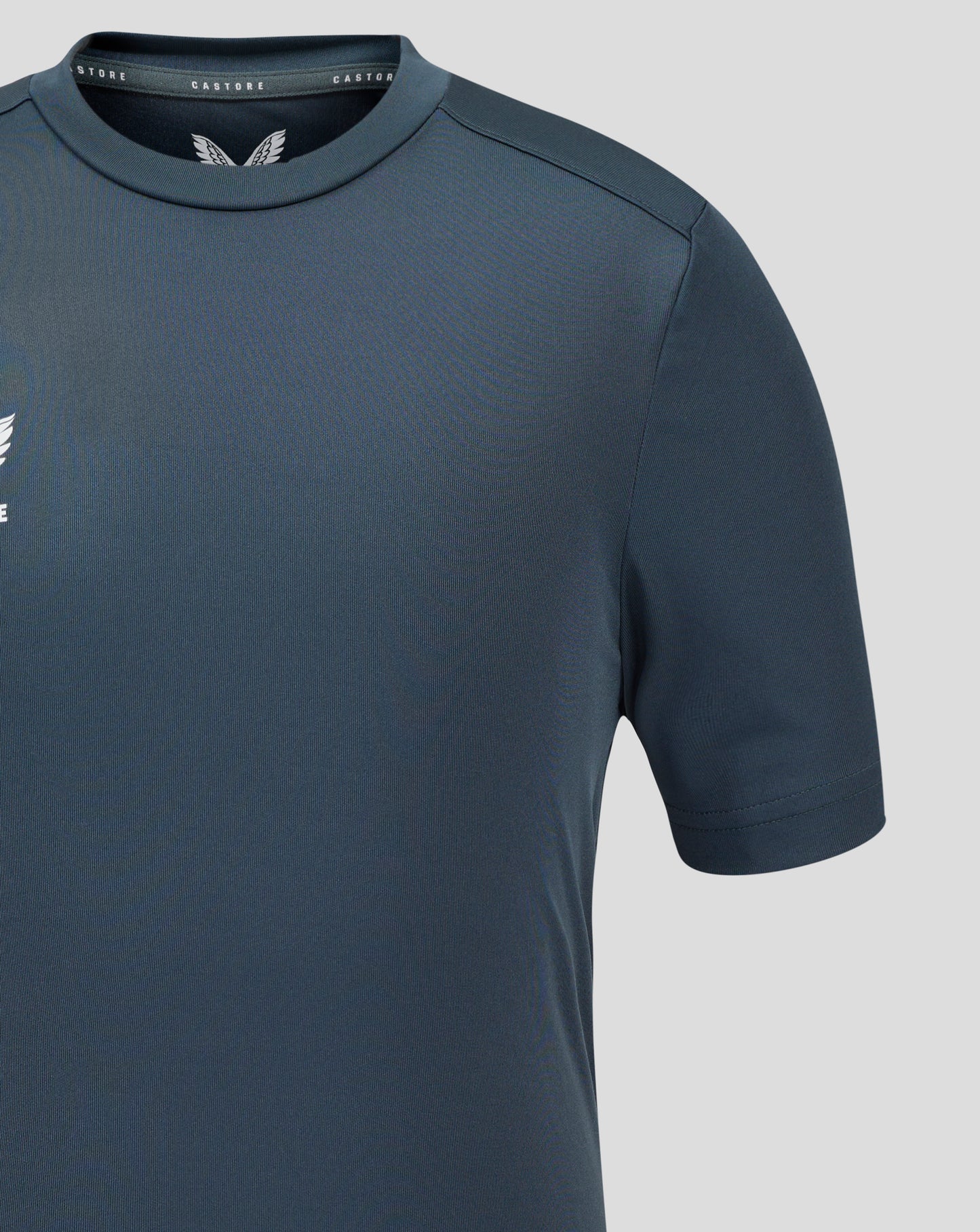 Close-up of dark slate Junior Performance Tee showing crew neck collar, set-in sleeve seam, and Castore branding near the neckline.