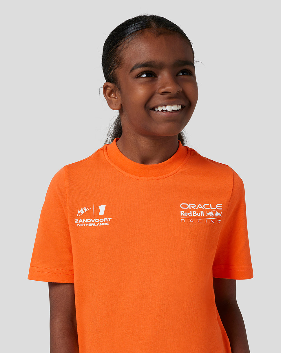 Smiling child wearing a bright orange Oracle Red Bull Racing Junior T Shirt with Verstappen graphic on the chest and team logos