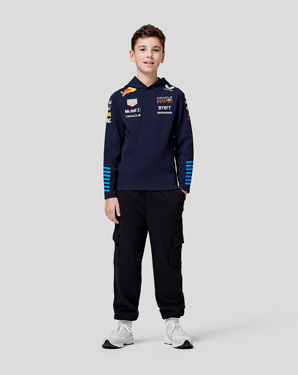 Front view of a young racer wearing navy teamline hoodie with chest logos blue striped sleeves black cargo pants and white sneakers.