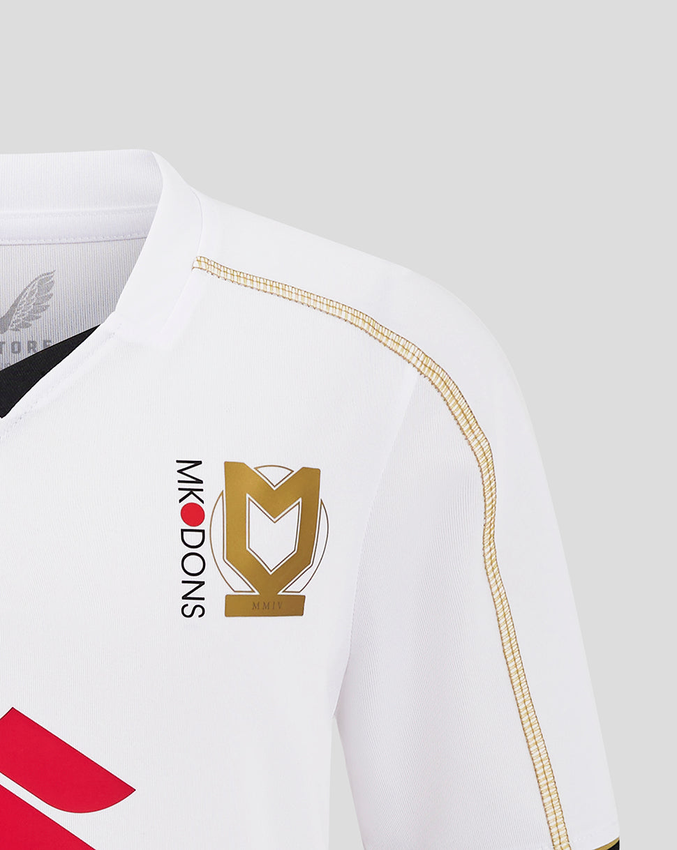 Close up of white MK Dons junior home shirt chest featuring a gold crest and vertical MK Dons text