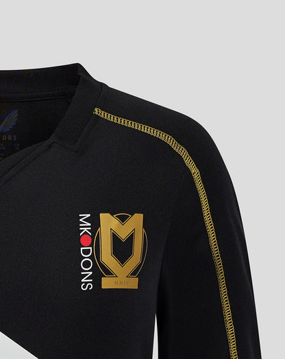 Close-up of chest on black MK DONS junior 24/25 third shirt showing a large gold crest and vertical MK DONS lettering beside it with gold shoulder seam.
