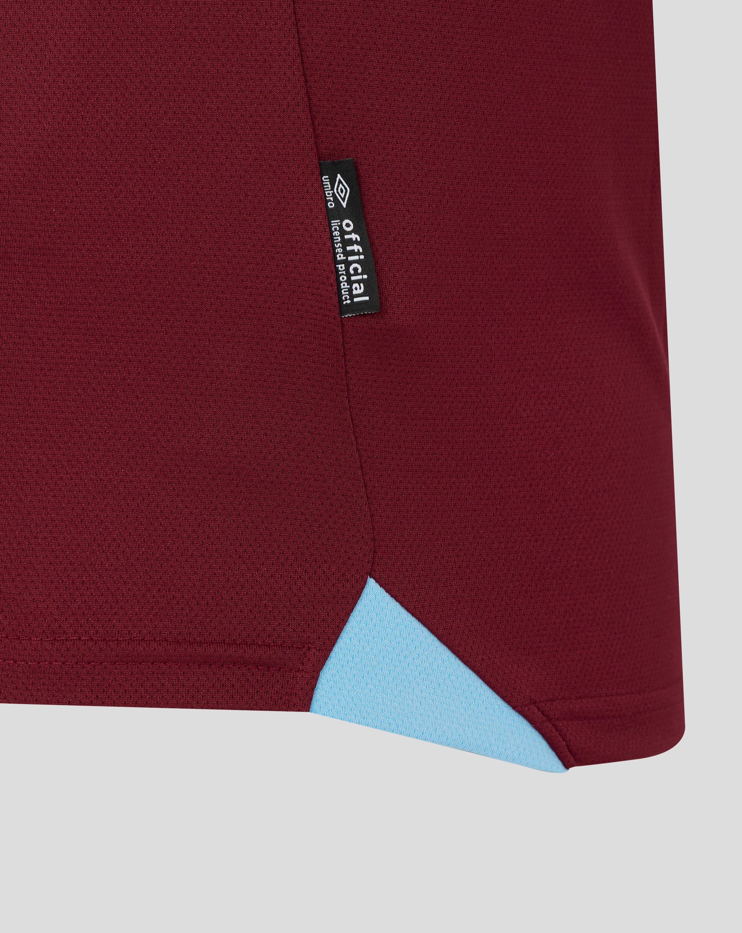 Close up of maroon fabric from a West Ham home shirt with a vertical black tag showing Official Licensed Product and the Umbro logo along the seam
