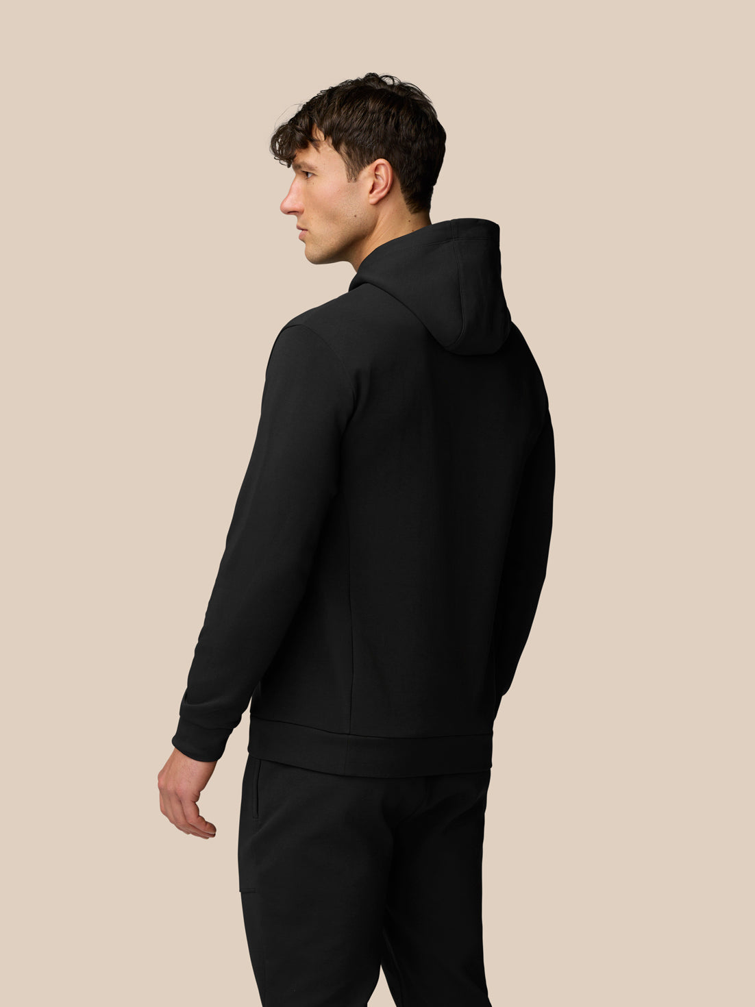 Side profile of a man wearing a black Travel Logo Hoodie against a beige background.
