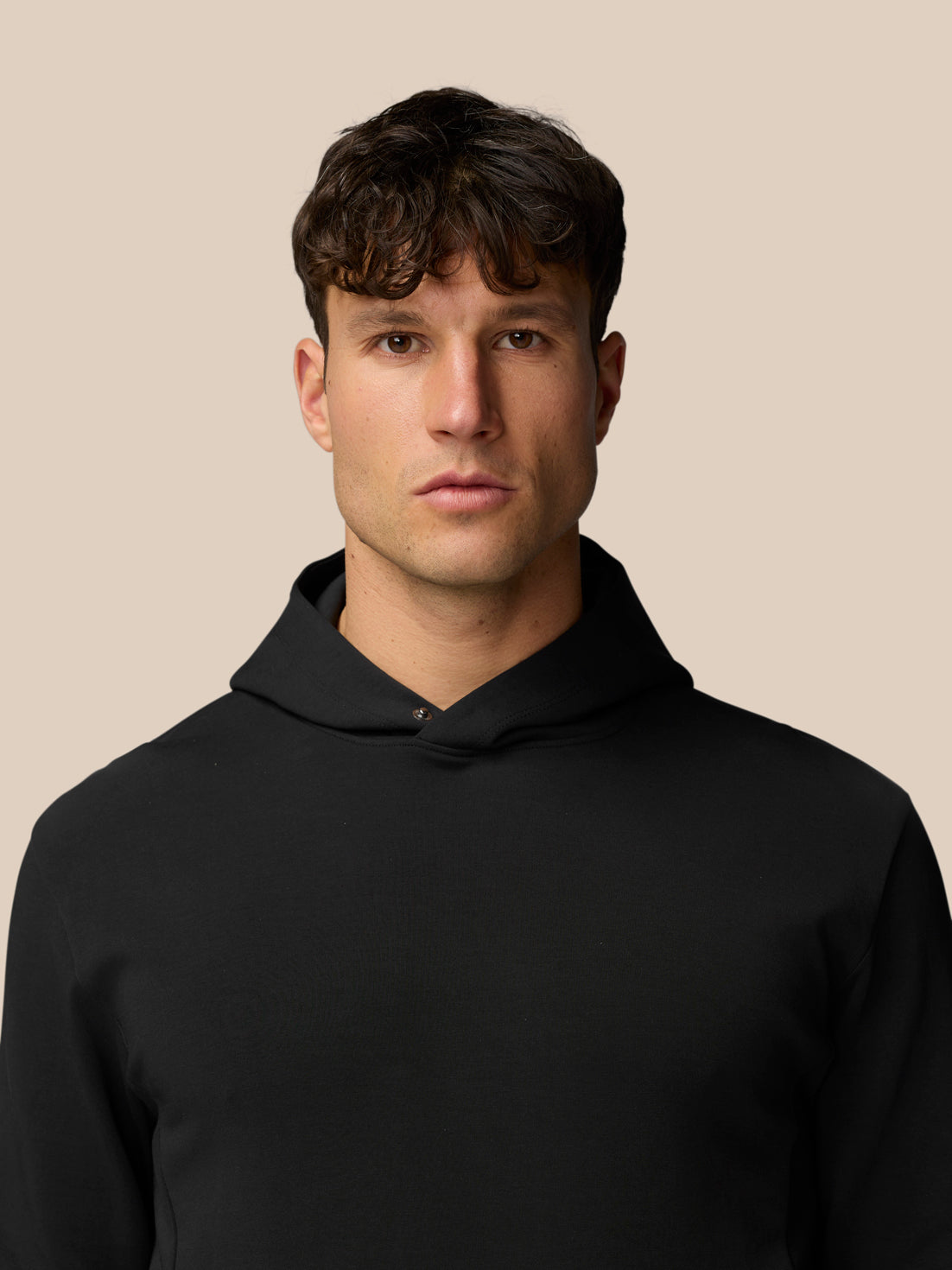 Front view of a man in a black Travel Logo Hoodie with the hood up against a beige backdrop.