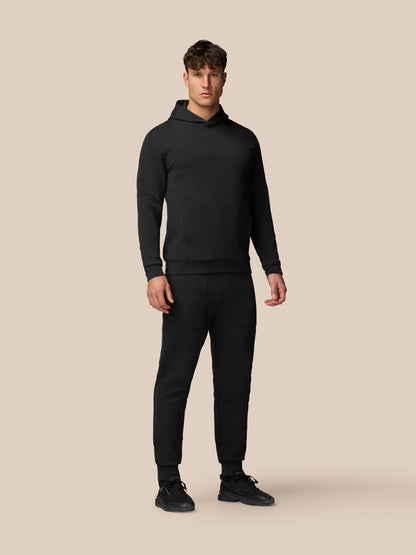 Male model wearing black Flex Travel Joggers and matching hoodie against a beige backdrop.