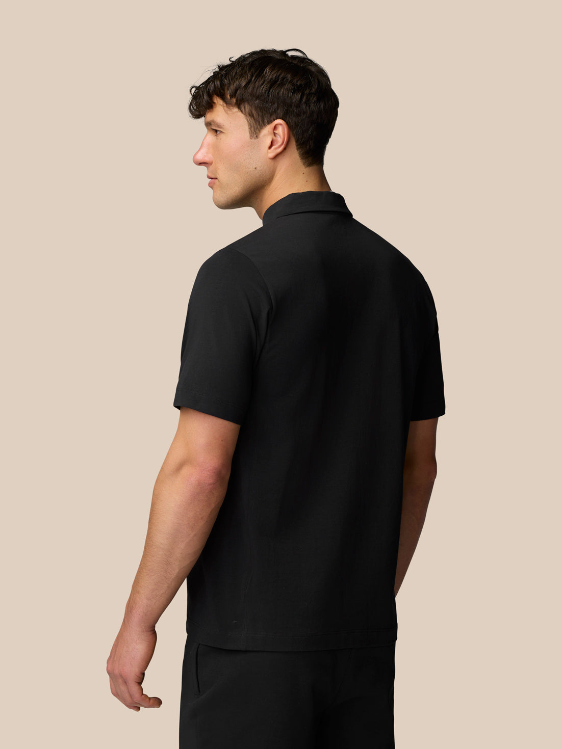 Man in black Travel Polo viewed from the side and back against a beige background
