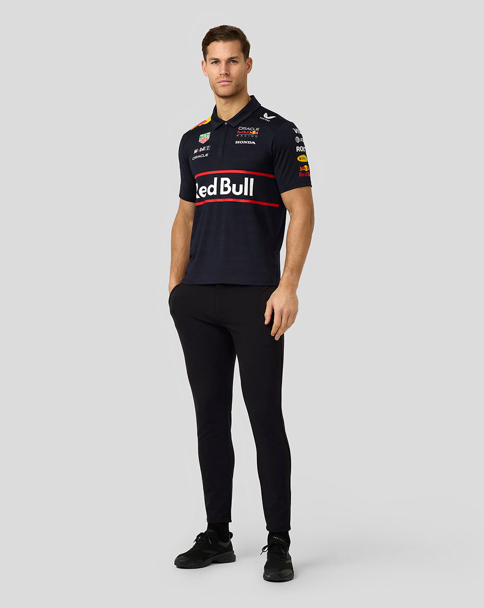 Full length view of a male model wearing a navy Red Bull Racing polo with sponsor logos and black pants.