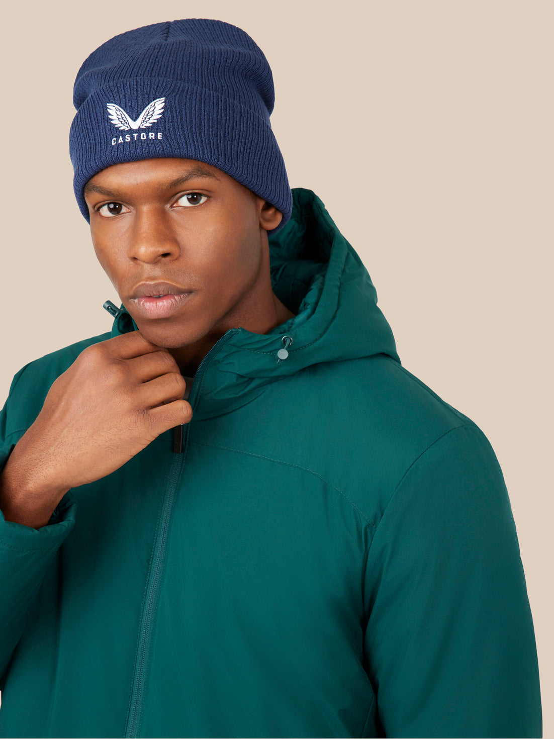 Male model wearing navy knit Active Beanie with white Castore logo and teal jacket against a beige background
