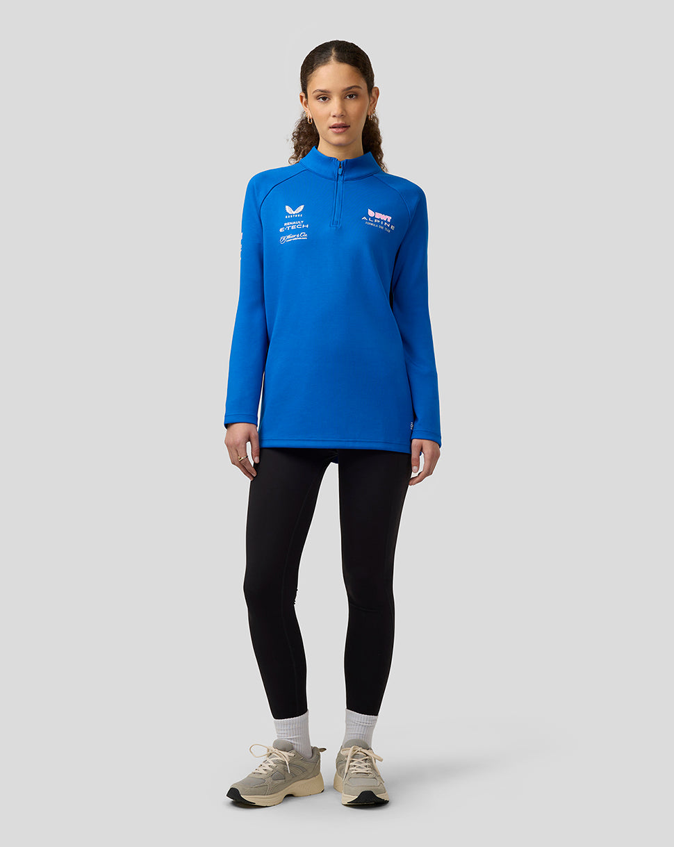 Woman wearing lapis blue BWT Alpine funnel neck quarter zip midlayer with team logos, paired with black leggings.