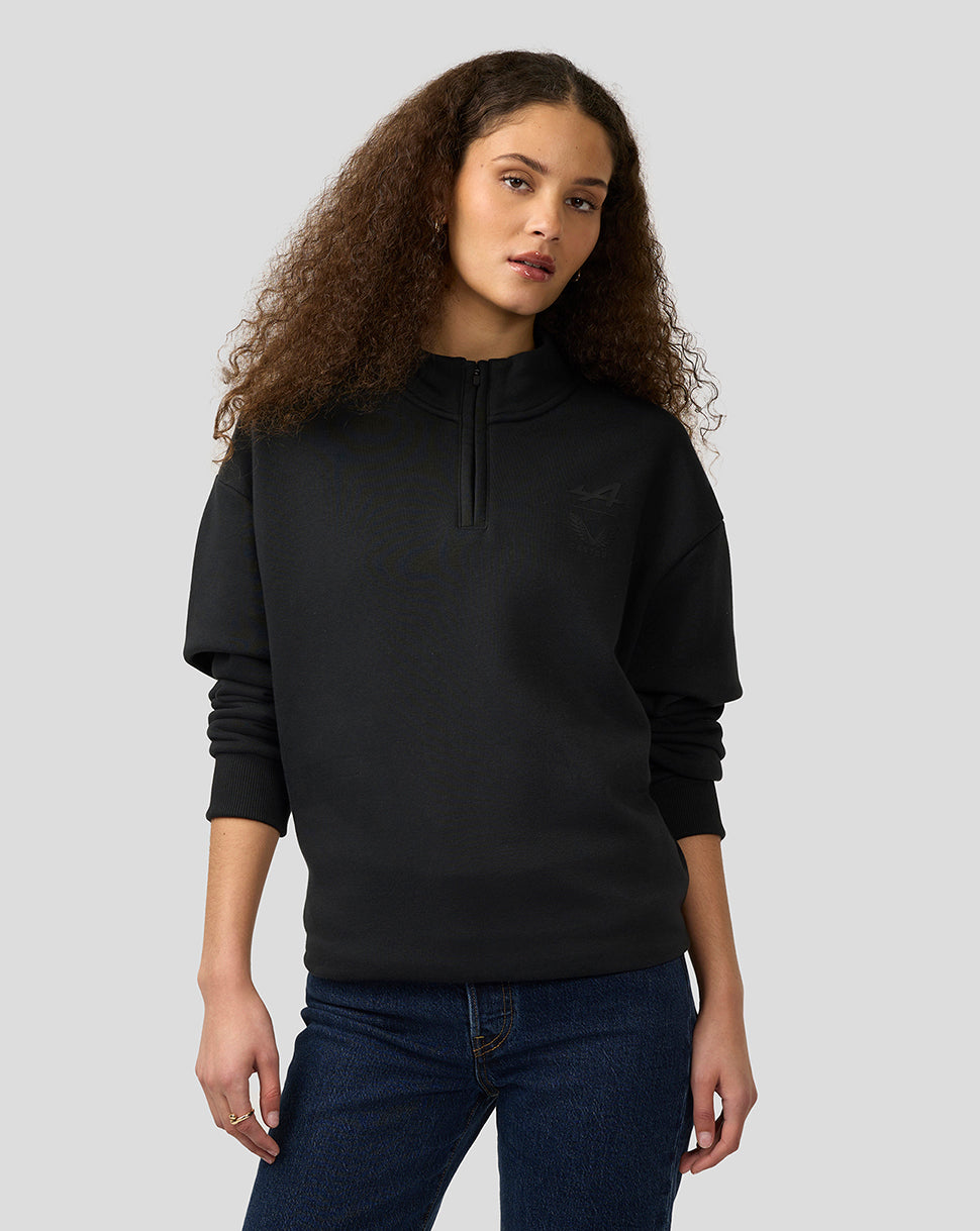 Female model in black Alpine Castore quarter zip pullover with relaxed fit standing against a light gray background
