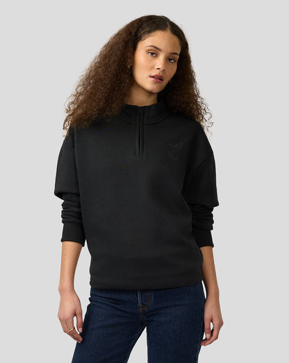 Female model in black Alpine Castore quarter zip pullover with relaxed fit standing against a light gray background
