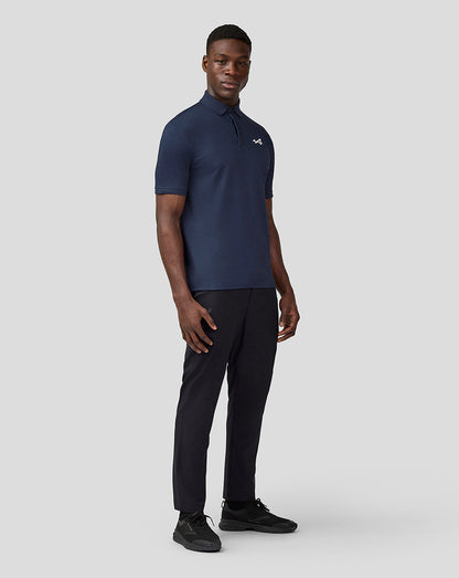 Male model in navy Alpine Core Essentials Polo Shirt with chest logo, paired with black trousers and sneakers.