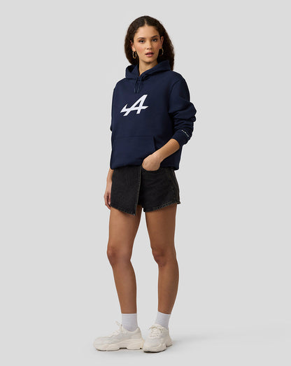 Female model in navy Alpine Core Essentials Hoodie with bold chest logo, paired with dark denim shorts and white sneakers