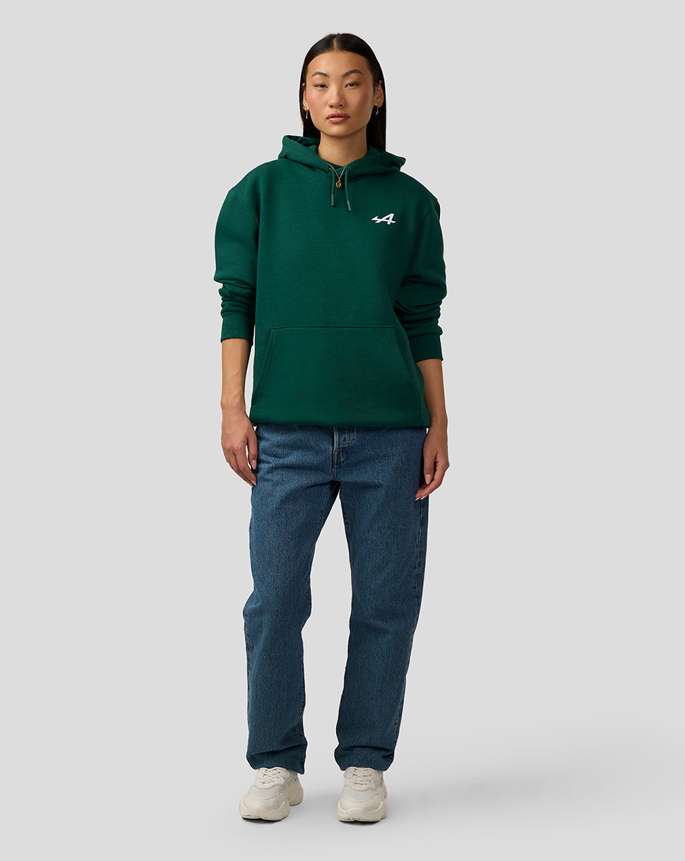 Female model wearing botanical green Alpine hoodie with white logo on chest, standing in casual jeans.